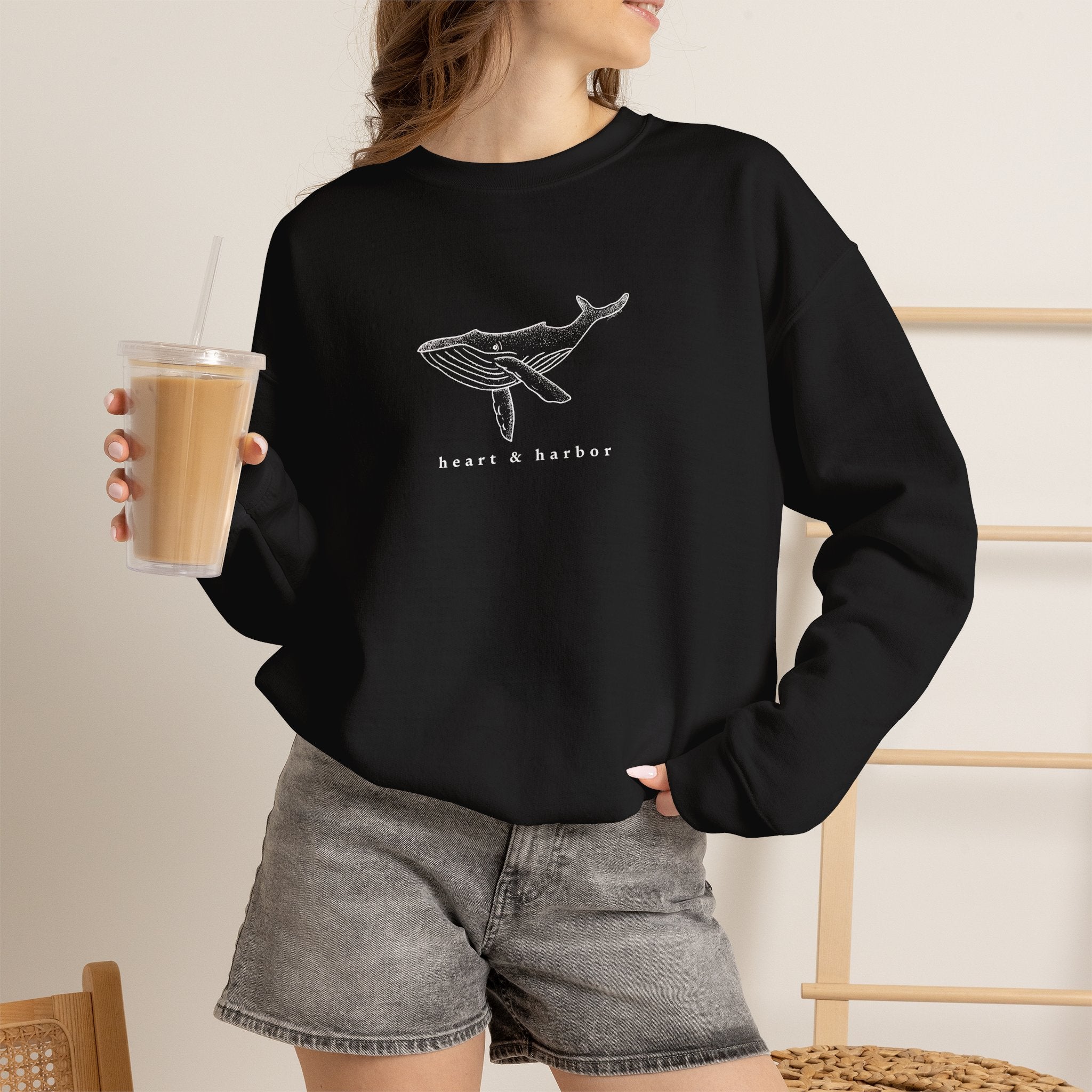 Person wearing a black hand-drawn whale crewneck sweatshirt by Heart & Harbor